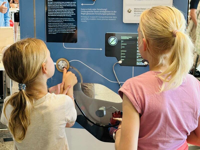 Zwei junge Mädchen interagieren mit einem Ausstellungsstück, drücken Knöpfe und lesen Informationen. Die Szene ist fesselnd und stellt das Lernen in den Mittelpunkt.