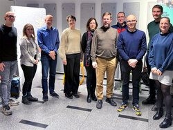 Eine Gruppe von zehn Personen steht in einem hellen Raum für ein Gruppenfoto zusammen. Im Hintergrund sind graue Akustikpaneele an der Wand zu sehen, und links steht eine Flipchart. Die Personen sind unterschiedlich gekleidet und lächeln in die Kamera.