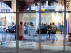 A group of people are standing in a room and interacting with each other. The view is from outside through a big window.