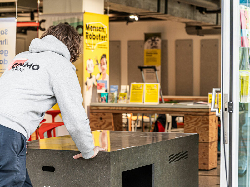 Person mit grauem COSMO-Hoodie schiebt eine schwarze Kiste in den Ausstellungsraum der Ausstellung 'Mensch, Roboter!' im COSMO Wissenschaftsforum.